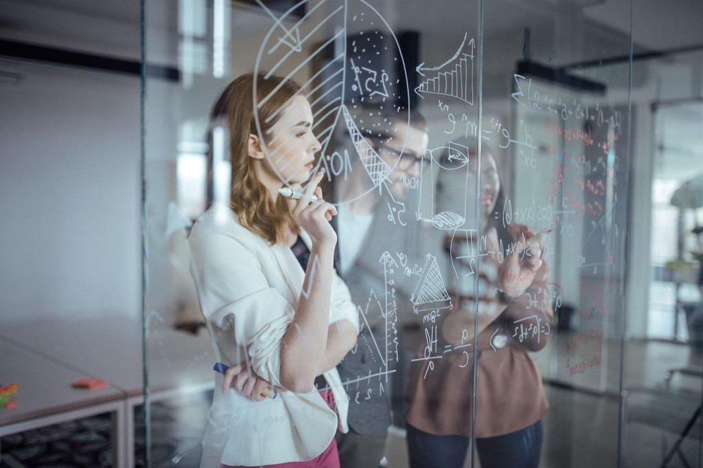 Three professionals collaborating at a glass wall with charts, graphs, and formulas